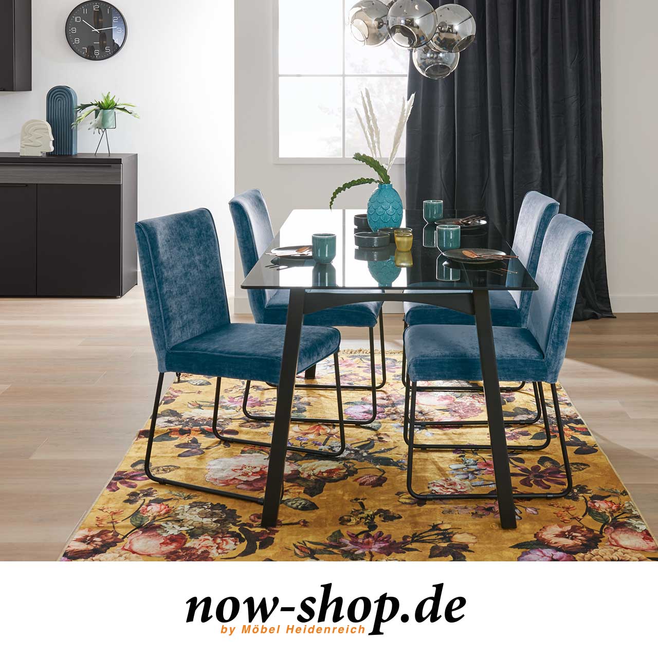 Esstisch mit getönter Glasplatte und schwarz lackiertem Gestell. Milieubild mit blauen samtig bezogenen Stühlen und Dekoration.