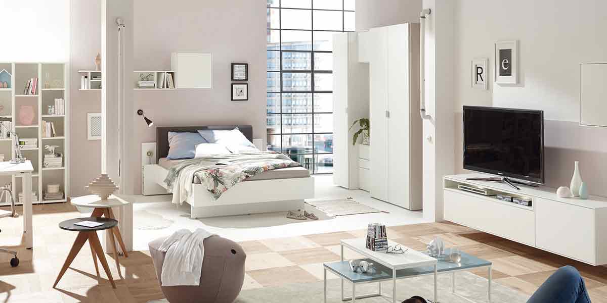 Titelbild Schlafzimmer Milieubild eines Schlafzimmers. In der Mitte ein Bett der Serie Basic, rechts ein Kleiderschrank und ein Sideboard mit Fernseher.
